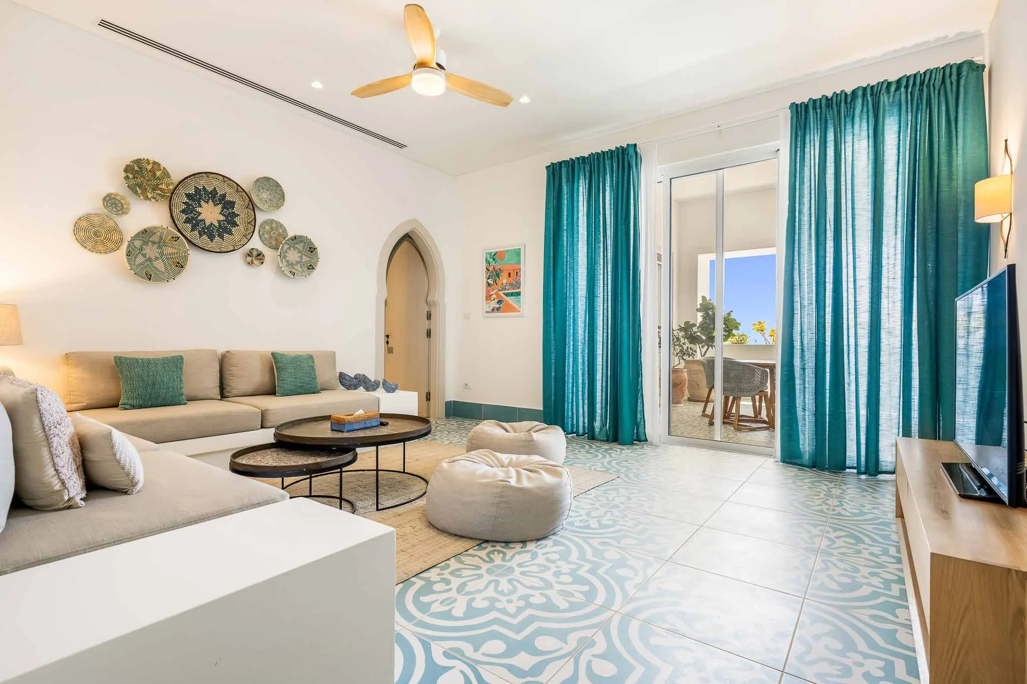 Living room with beige sectional sofa, green pillows, round black coffee tables, white and blue patterned floor tiles, turquoise curtains, a sliding glass door leading to an outdoor patio with potted plants, wall art, ceiling fan with wooden blades, and a TV on a wooden stand.