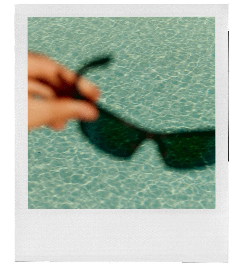A blurry image of a hand holding a pair of black sunglasses over a swimming pool with clear blue water.