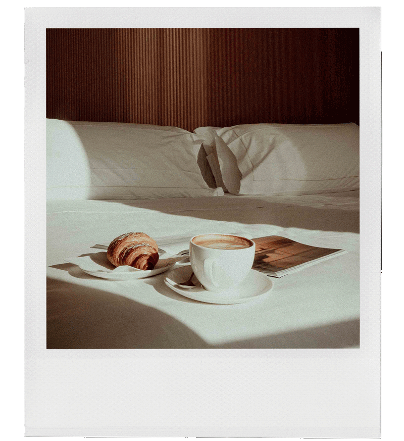 A breakfast tray with a croissant and a cup of coffee on a white bed in a well-lit bedroom.