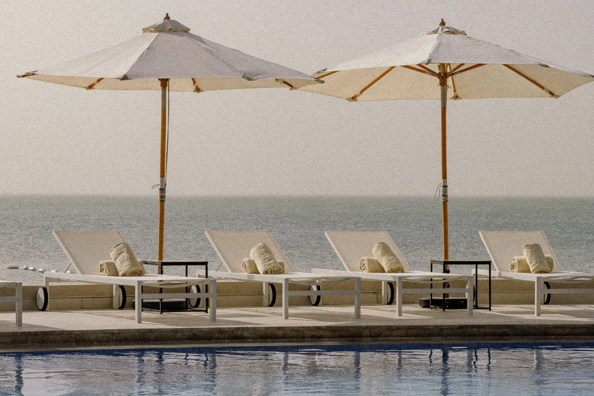Beach scene with two white beach umbrellas, lounge chairs with rolled towels, by a pool with ocean in the background.