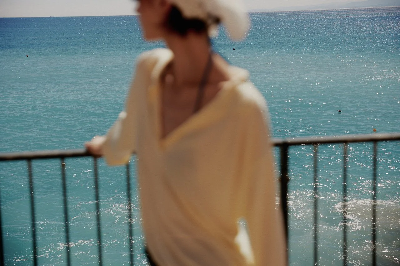 Blurred woman in a wide-brim hat and light-colored top standing by a seaside railing with the ocean in the background.