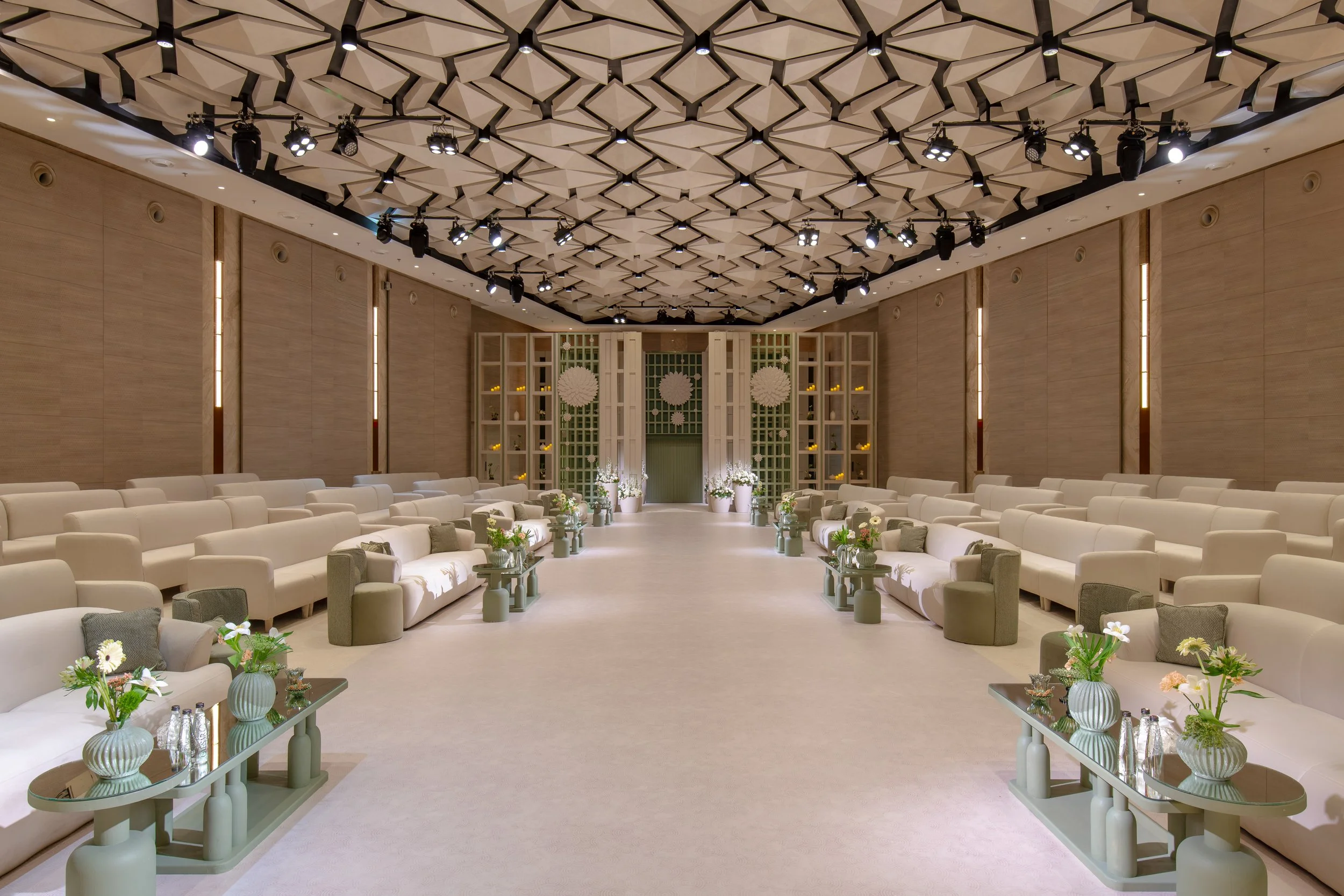 Elegant event hall with rows of white cushioned seating, light-colored wooden walls, geometric ceiling design with stage lighting, and floral arrangements on side tables.