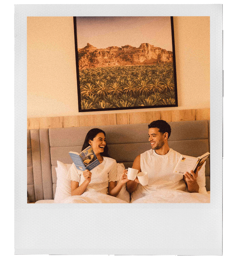 A young couple sitting up in bed, smiling and laughing. They are holding books and mugs, with a large landscape painting hanging on the wall behind them.