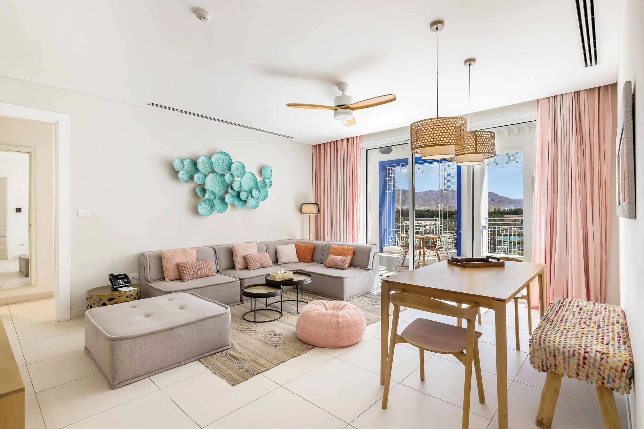Living room with beige sofa, pink cushions, marble coffee tables, beige ottoman, pink pouf, wooden dining table and chairs, pink curtains, wall art, floor lamp, ceiling fan, and large windows showing mountainous landscape outside.