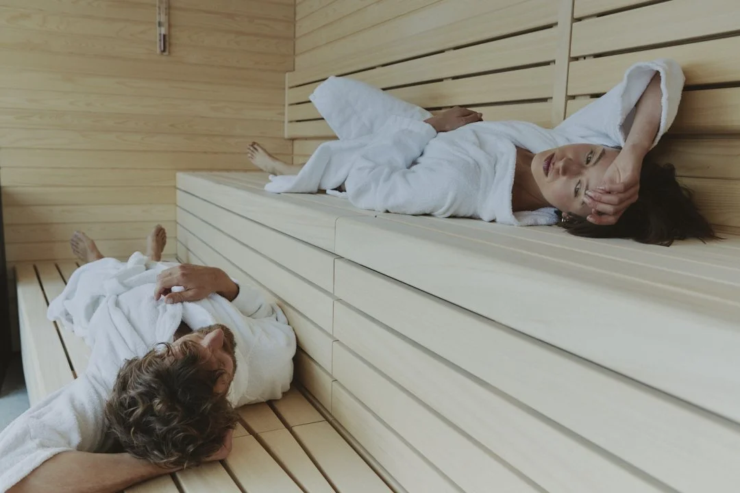 A woman and a man relaxing in a wooden sauna, both wearing white robes. The woman is lying on the bench with her head resting on her hand, and the man is lying on the floor with his head on the bench, both looking relaxed.