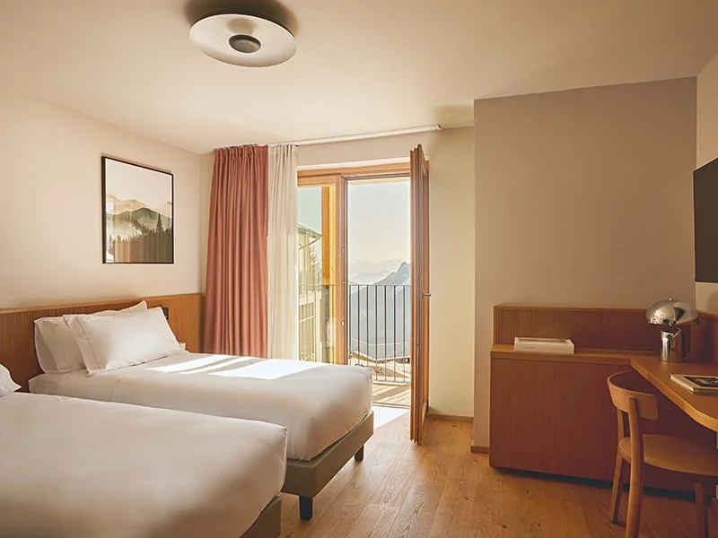 A hotel room with two beds, a balcony door with pink and white curtains, a framed landscape picture on the wall, a wooden desk with a lamp, and a chair.