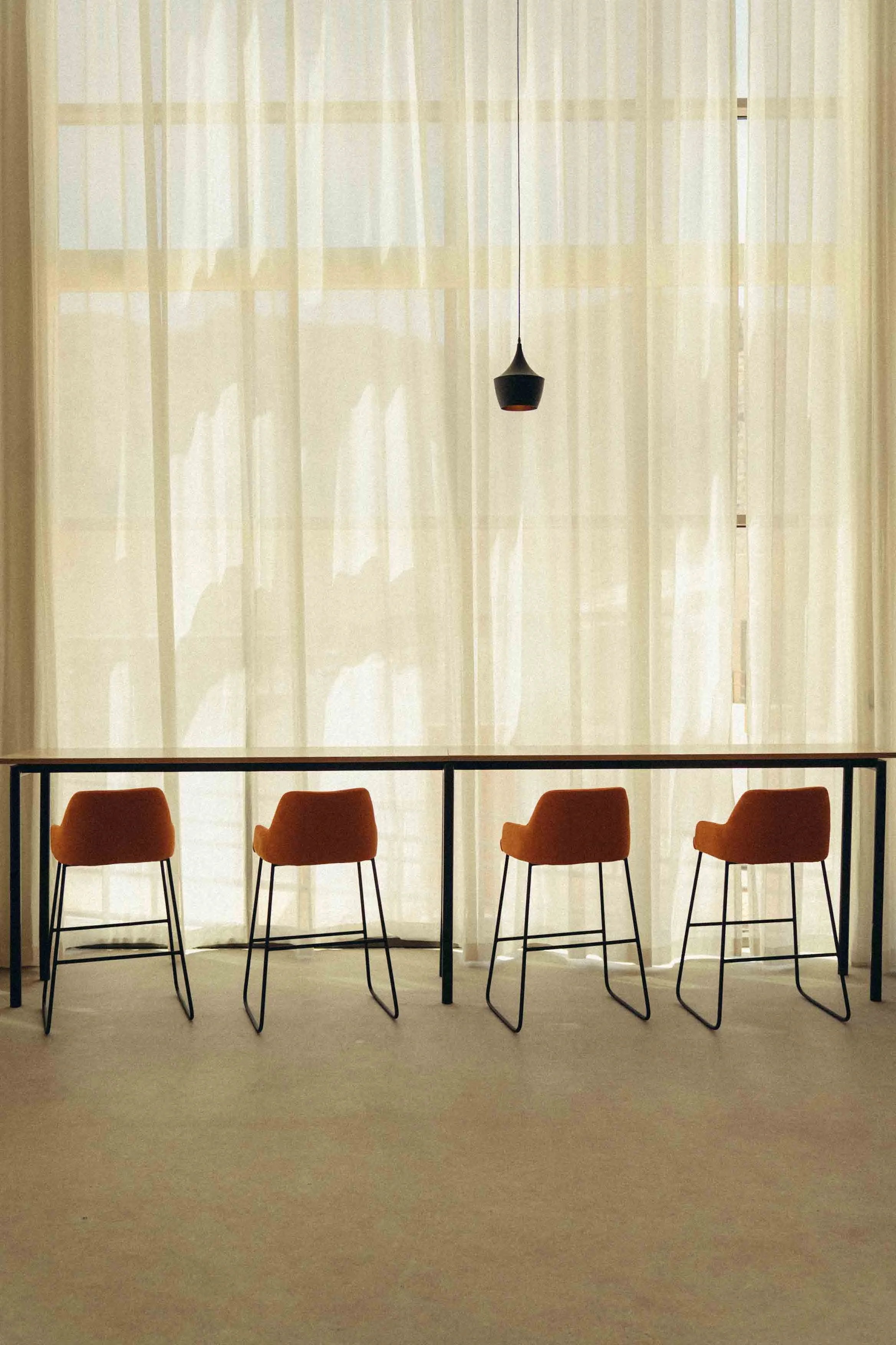 An interior room with a row of five orange barstools with black legs lined up along a black countertop. Behind the curtains, there is a window letting in natural light. A black pendant lamp hangs from the ceiling.