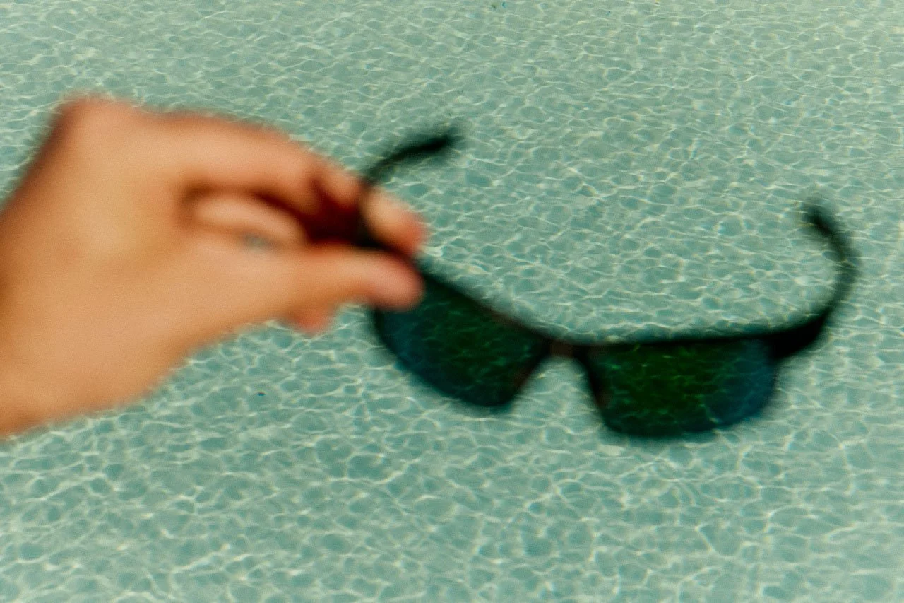 A hand holding a pair of green sunglasses above a swimming pool with tile water surface.