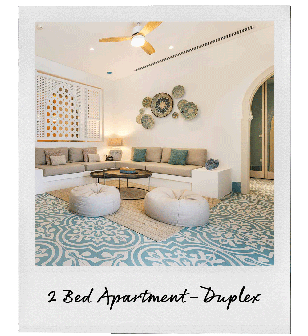 Living room in a 2-bedroom duplex apartment with beige couches, blue and beige pillows, a round coffee table, two white poufs, and decorative wall art, featuring a ceiling fan and patterned floor tiles.