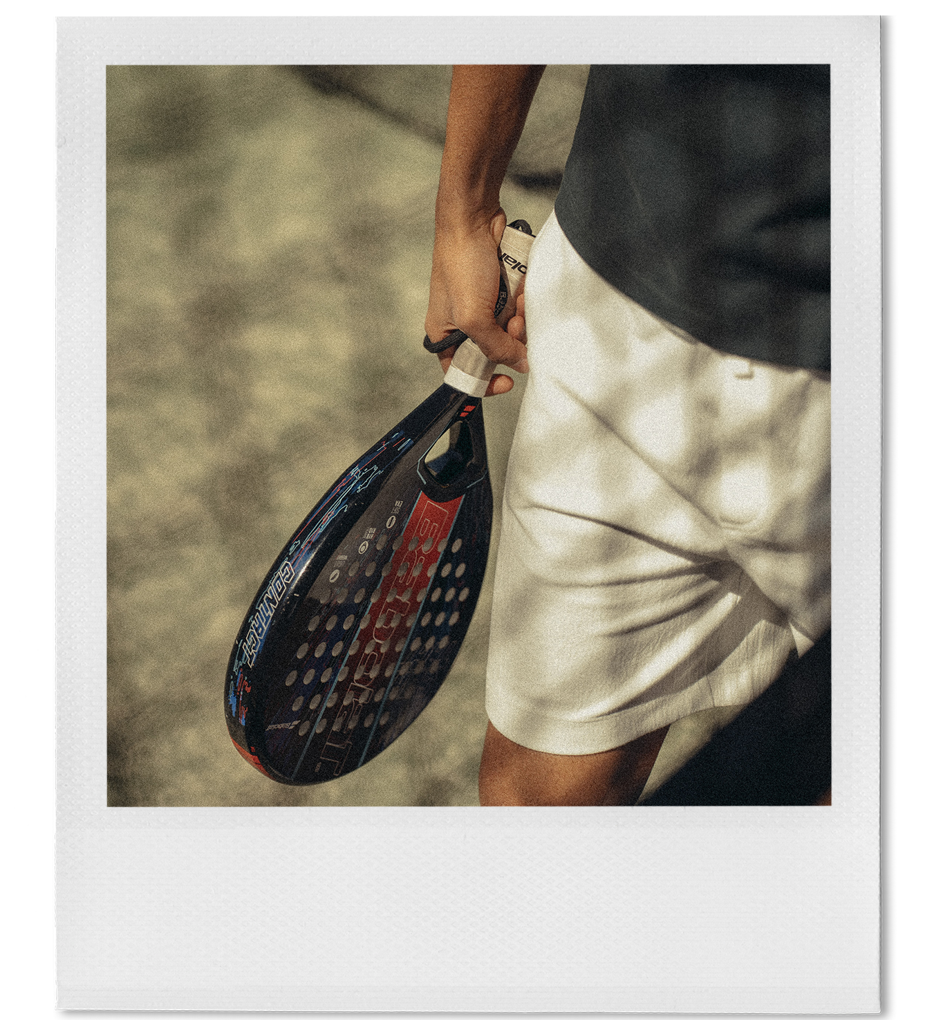 Close-up of a person holding a black and red padel racket, wearing white shorts and a dark shirt.