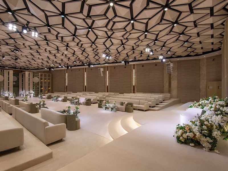 Elegant event hall with a high geometric ceiling, beige seating arranged in rows, floral centerpieces on small tables, and soft lighting for a formal occasion.