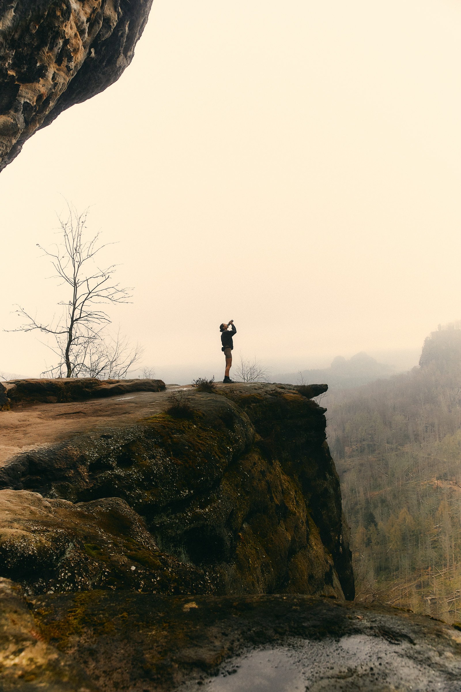 A person standing on a rocky ledge, taking a photo or looking through a camera, with a foggy landscape and leafless trees in the background.