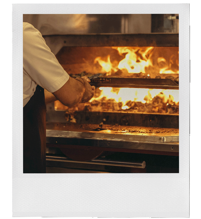 A chef in a white uniform and apron is using long tongs to handle food over a grill with open flames in a restaurant kitchen.
