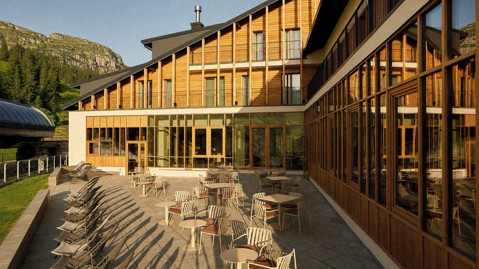 Outdoor patio area with chairs and tables in front of a modern multi-story building with large windows and wooden accents, set against a scenic mountain background.