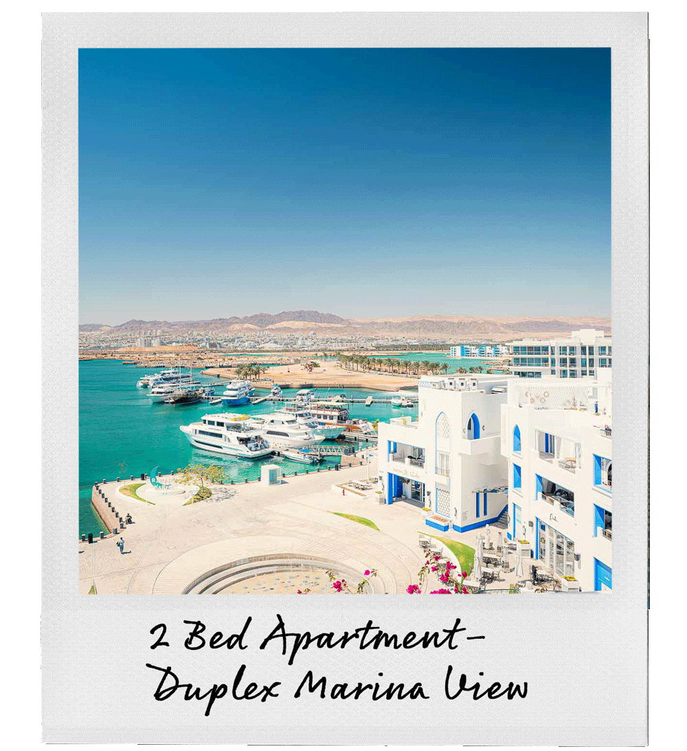View of a marina with yachts, white and blue buildings, and mountains in the background, under a clear blue sky.
