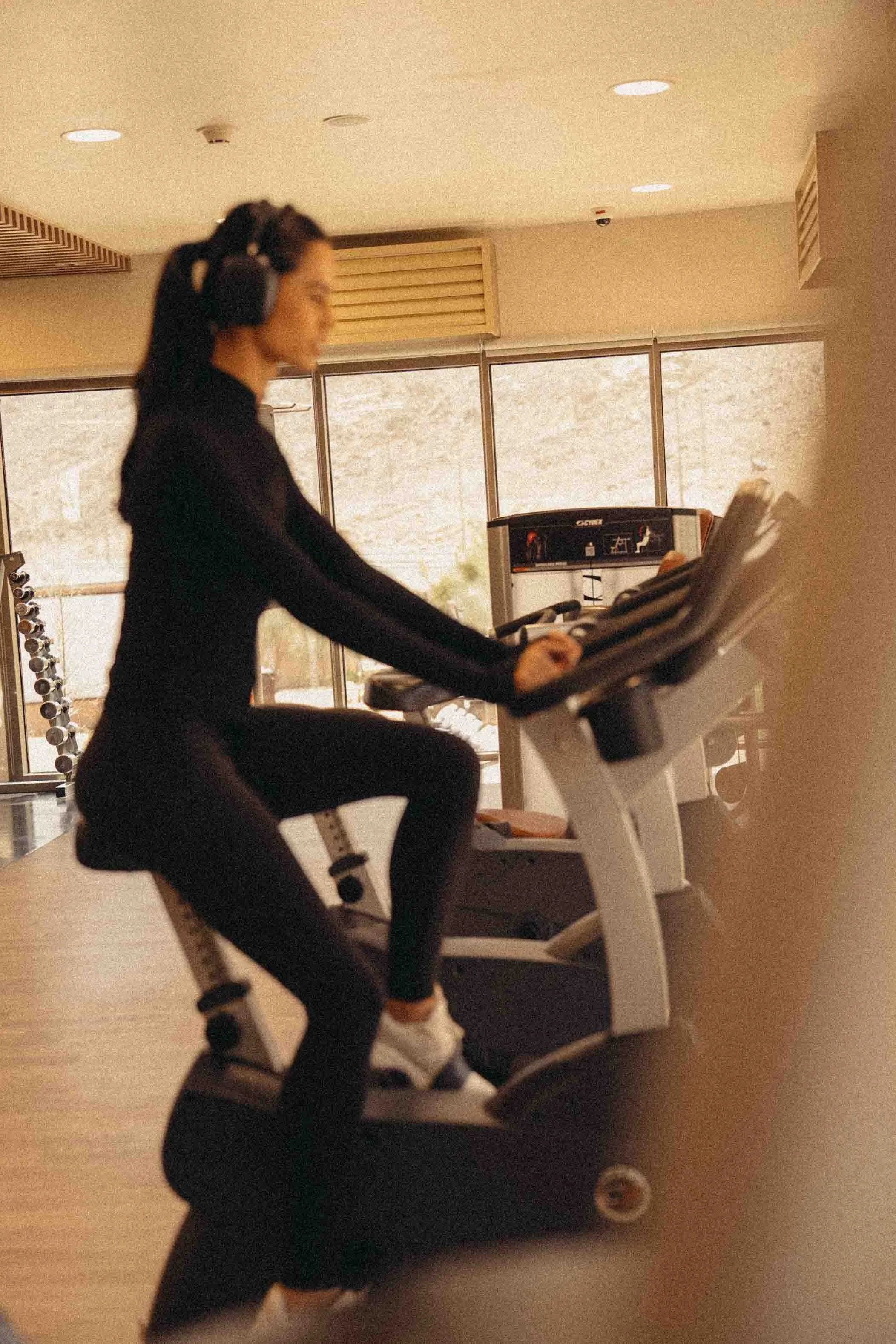 A woman with long dark hair in a ponytail, wearing headphones and activewear, sitting on a rowing machine in a gym with large windows and exercise equipment.