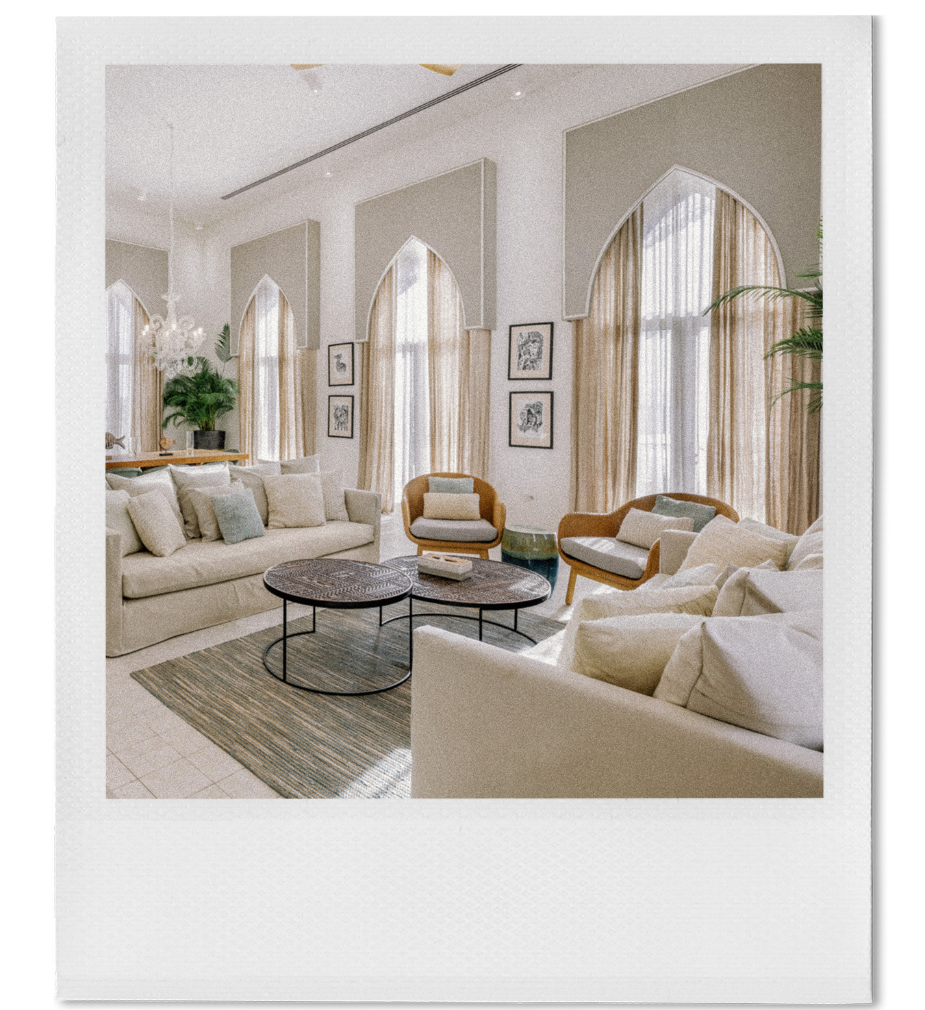 Living room with beige sofas, wooden coffee tables, and armchairs, large arched windows with sheer curtains, framed black and white artwork on the walls, potted plants, chandelier, and area rug.