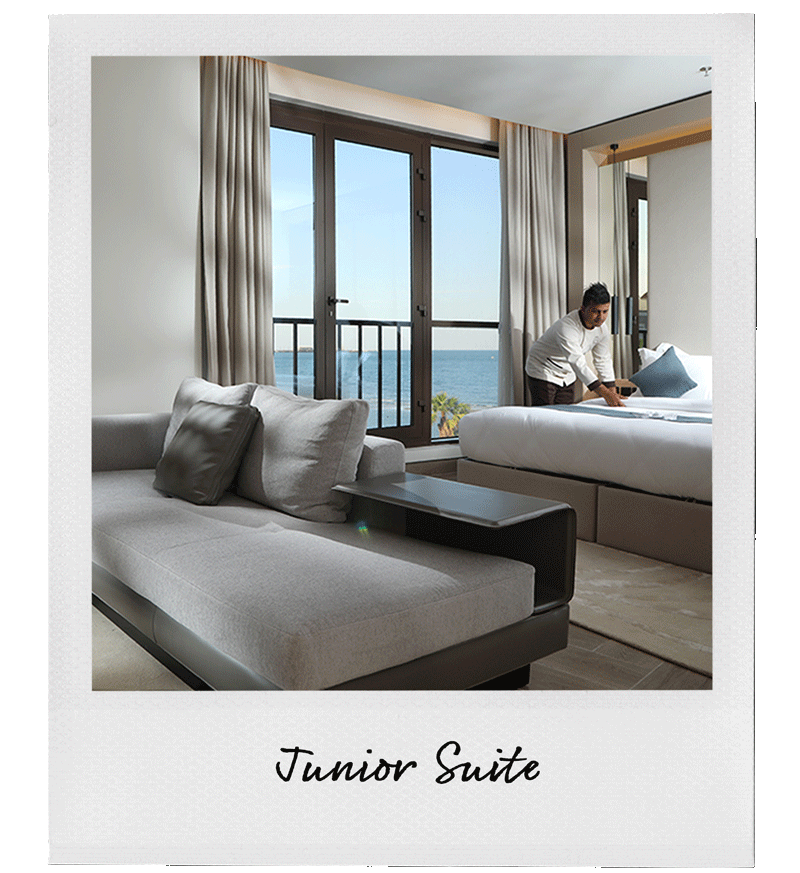 A hotel room with a large window view of the ocean, a gray sofa with pillows, a bed with person making the bed, and beach scenery outside.
