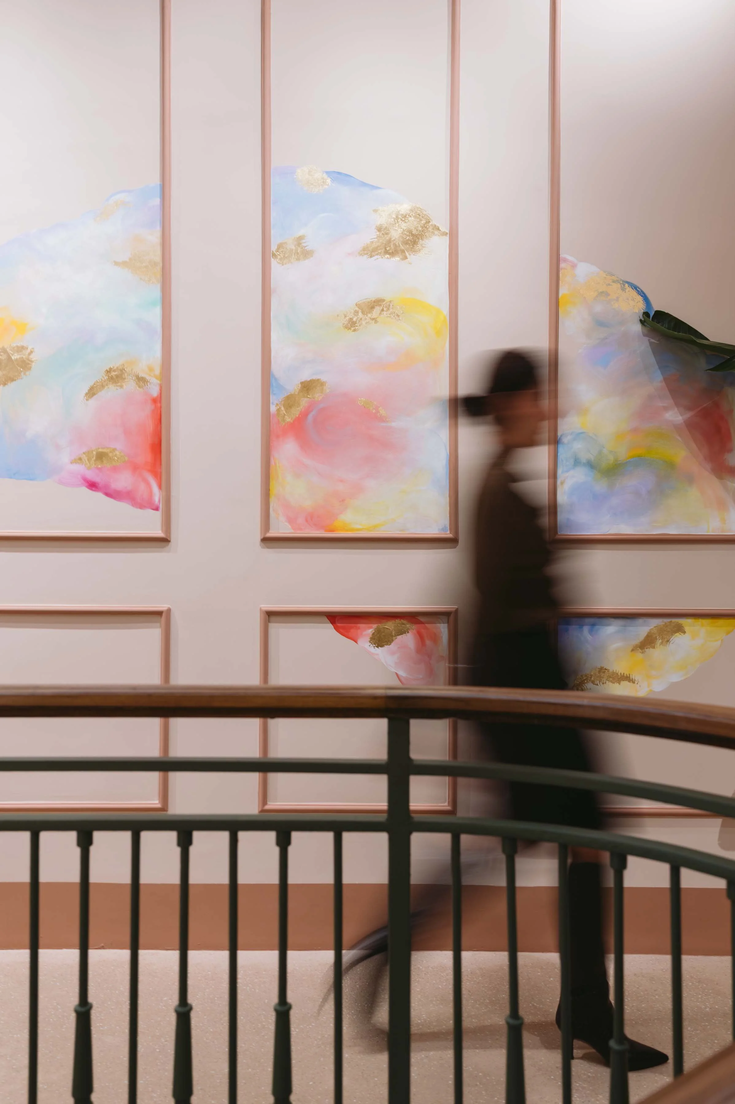 A person walking past a wall with abstract art in soft pastel colors and gold accents. The person appears blurred, and the photo is taken from behind a black railing.