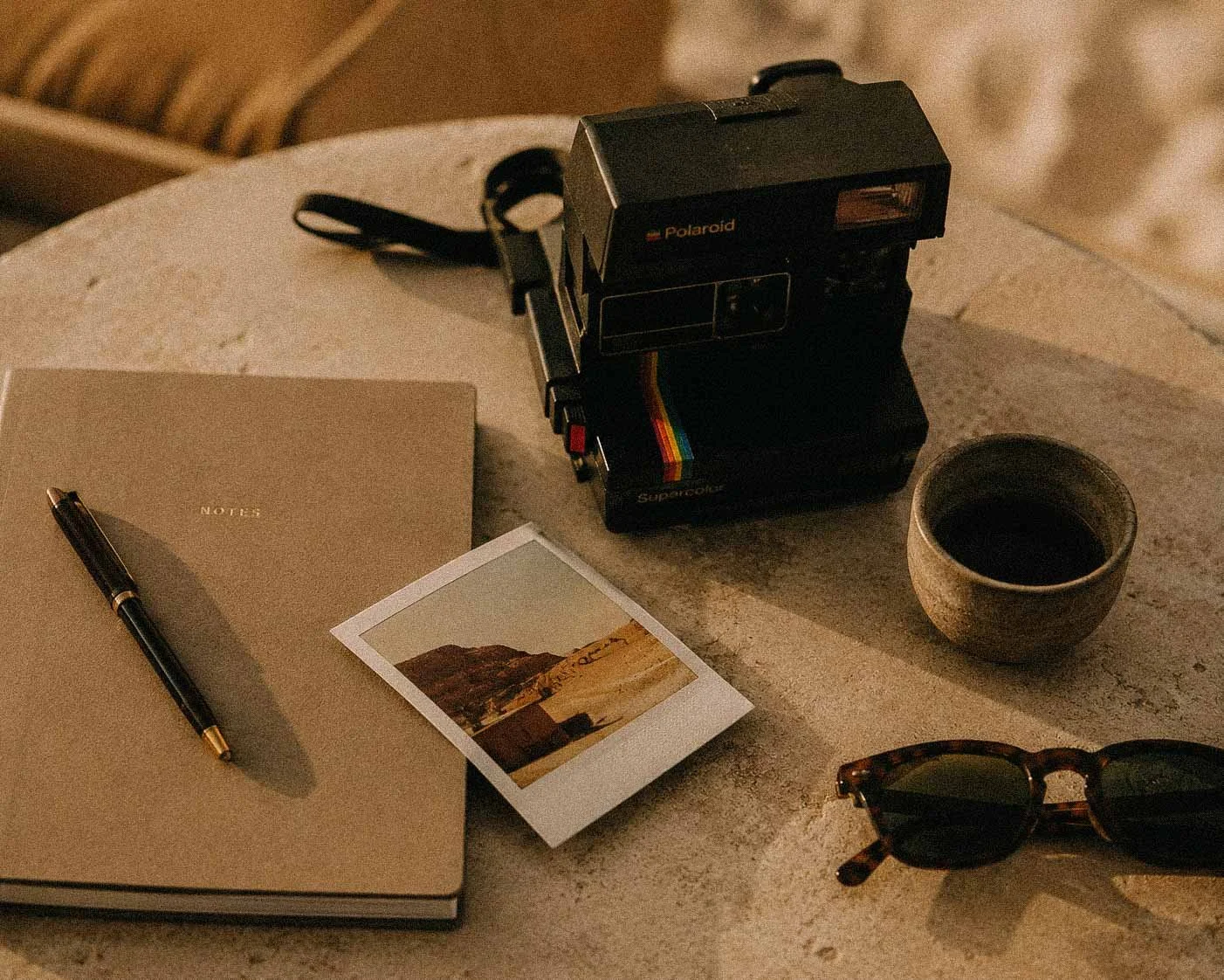 A table with a beige notebook, a black pen, a small instant photo, a polaroid camera, a coffee cup, and a pair of sunglasses.