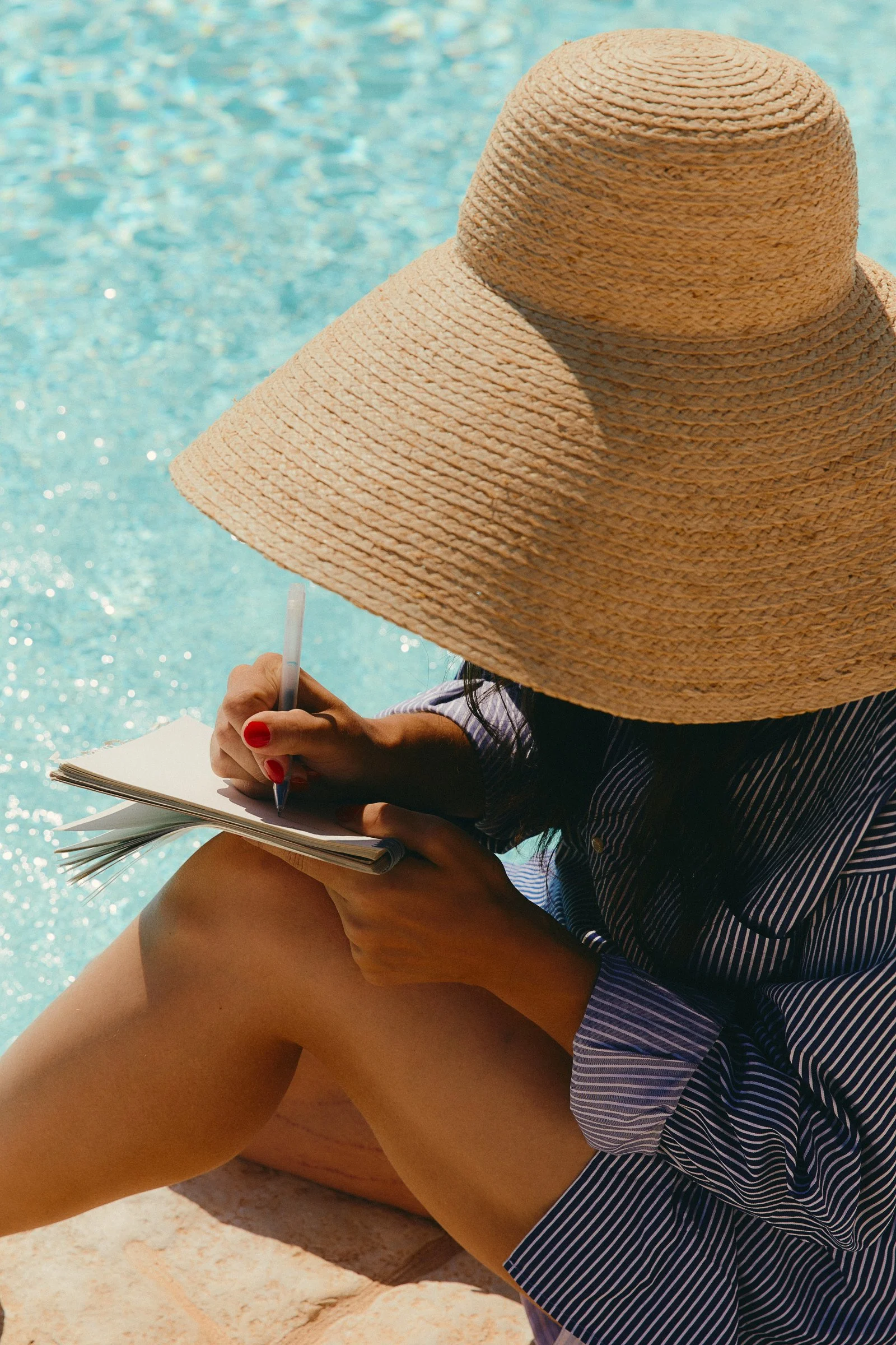 Woman sitting by a pool, wearing a large straw hat and a striped shirt, writing in a notebook.