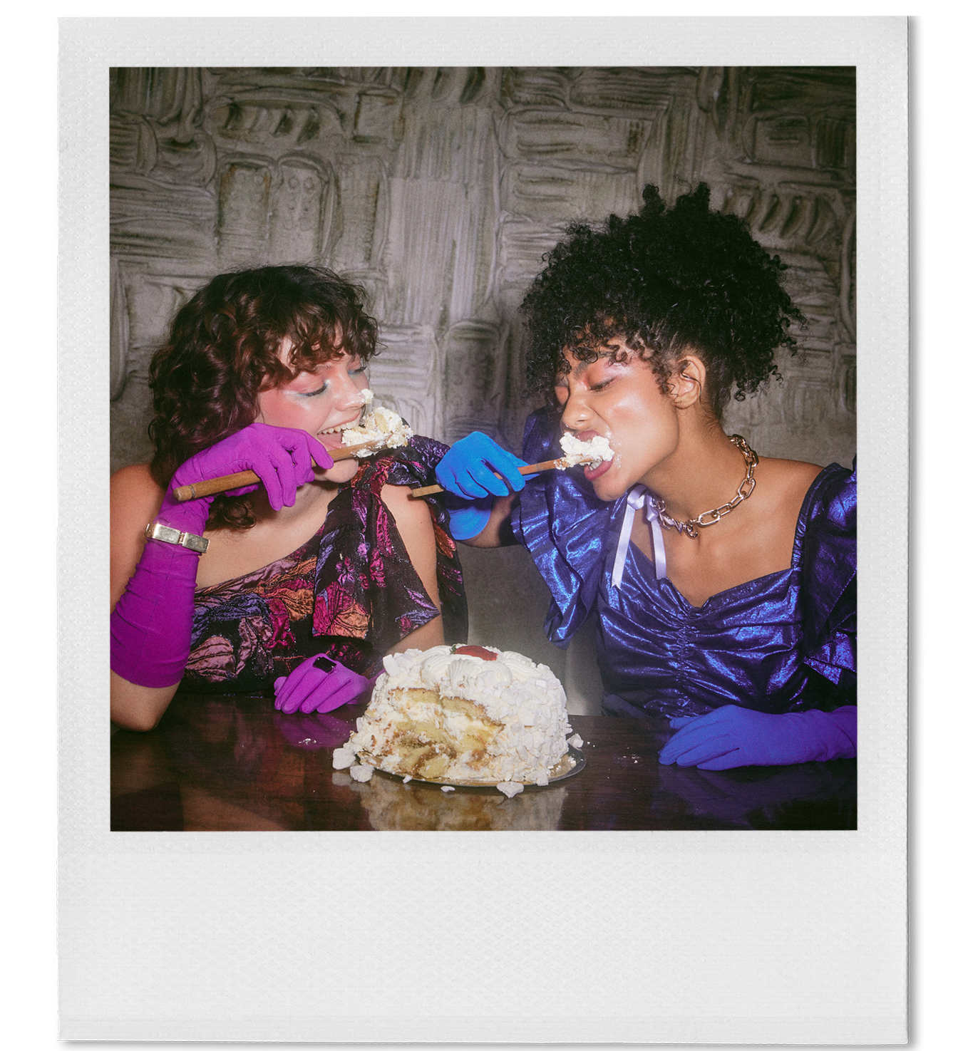 Two women with curly hair, one with brown and the other with black, wearing colorful dresses and gloves, sharing a slice of cake with whipped cream, using forks, in front of a sliced cake on a dark wooden table, against a textured gray wall.