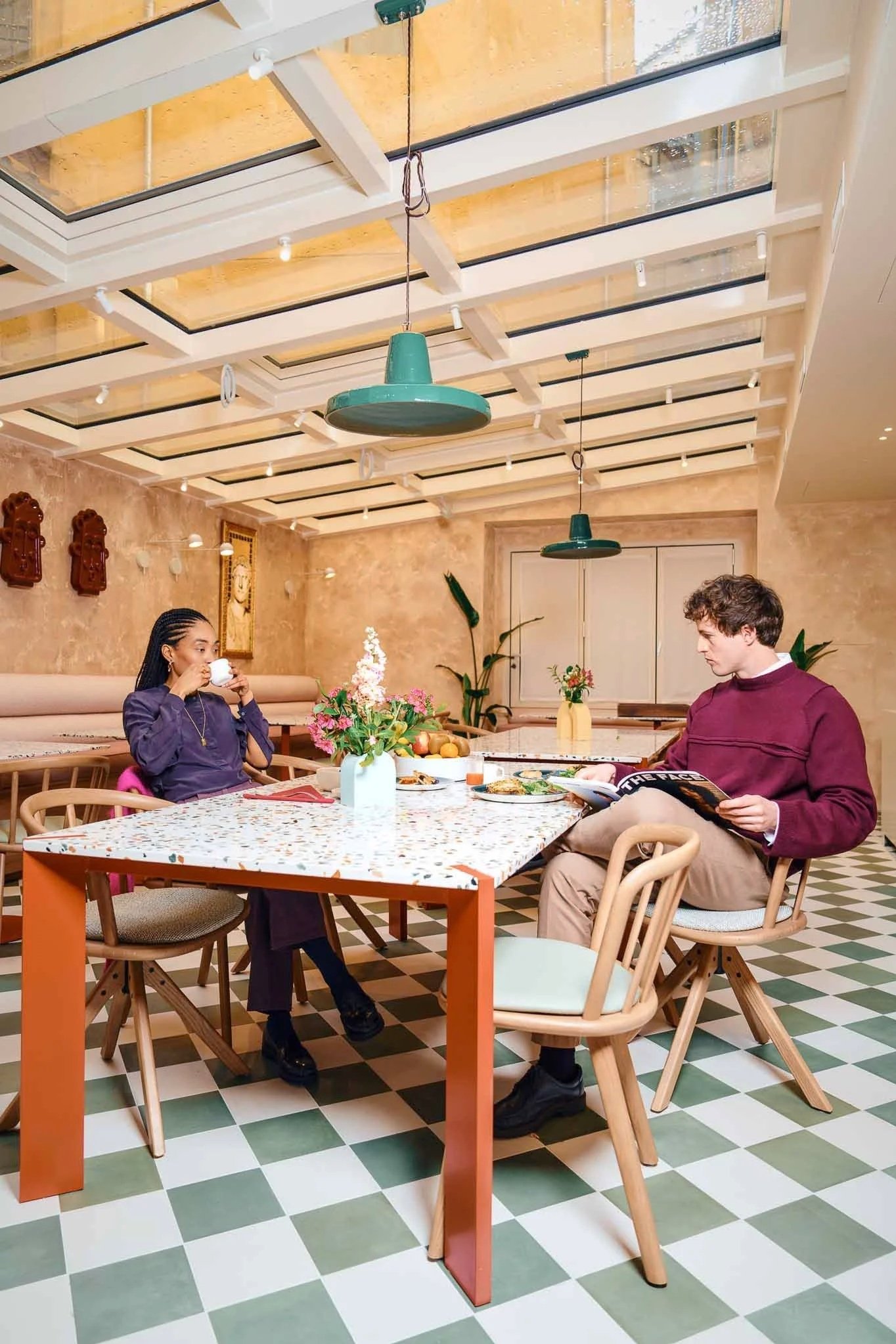 A woman with braided hair and a man with curly hair sitting at a colorful table in a cafe, with flowers and food on the table, and artwork on the tan walls.