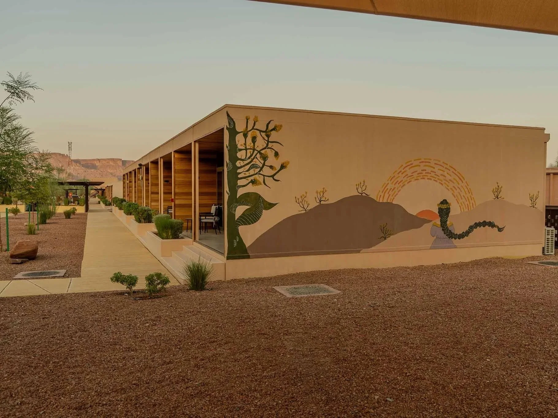 A building with a mural of a girl with a long braid in a desert landscape at sunset, featuring a large tree and mountains.