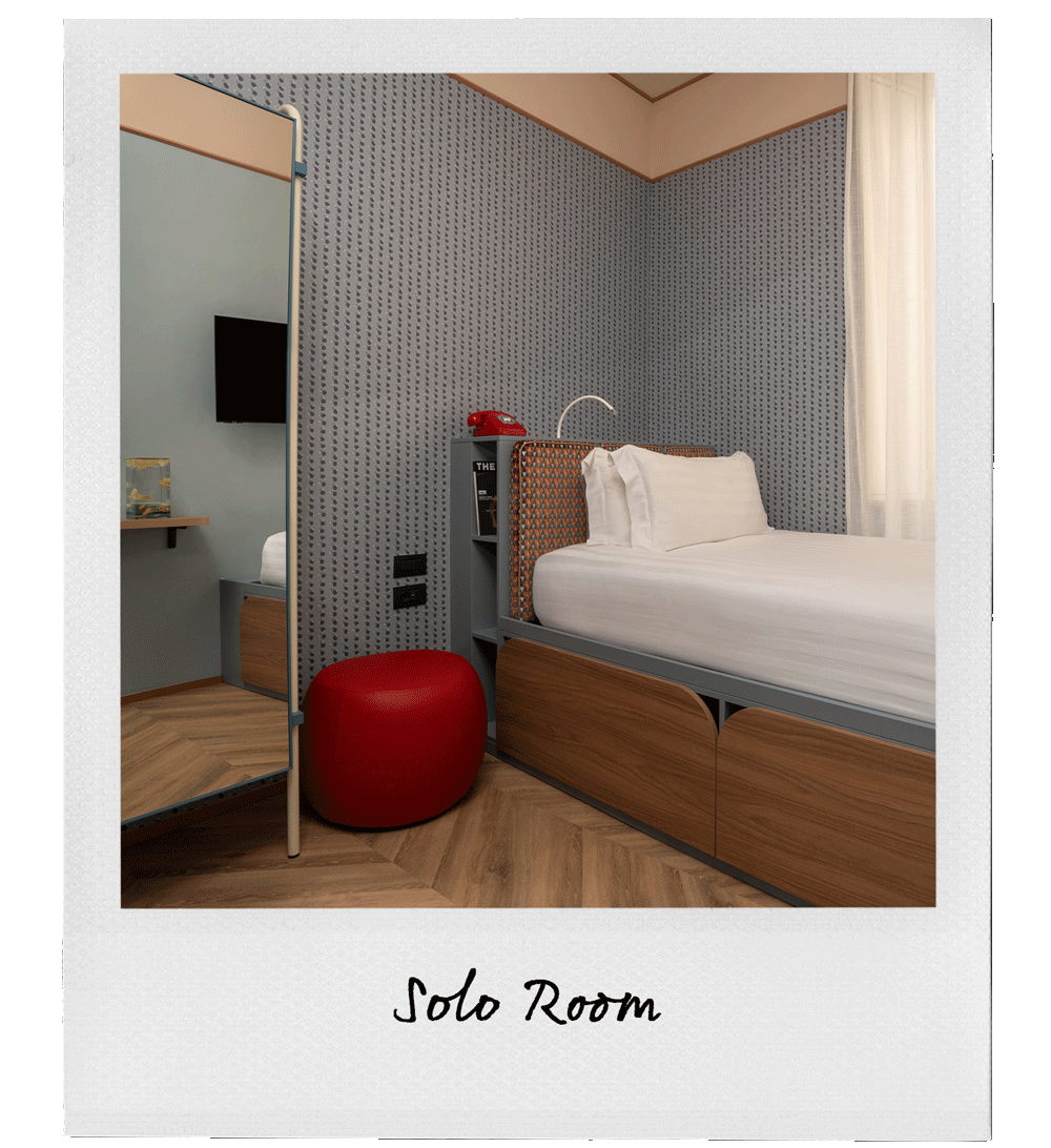 A modern hotel room with a bed, a red ottoman, a nightstand with a red vintage telephone, and a window with curtains.