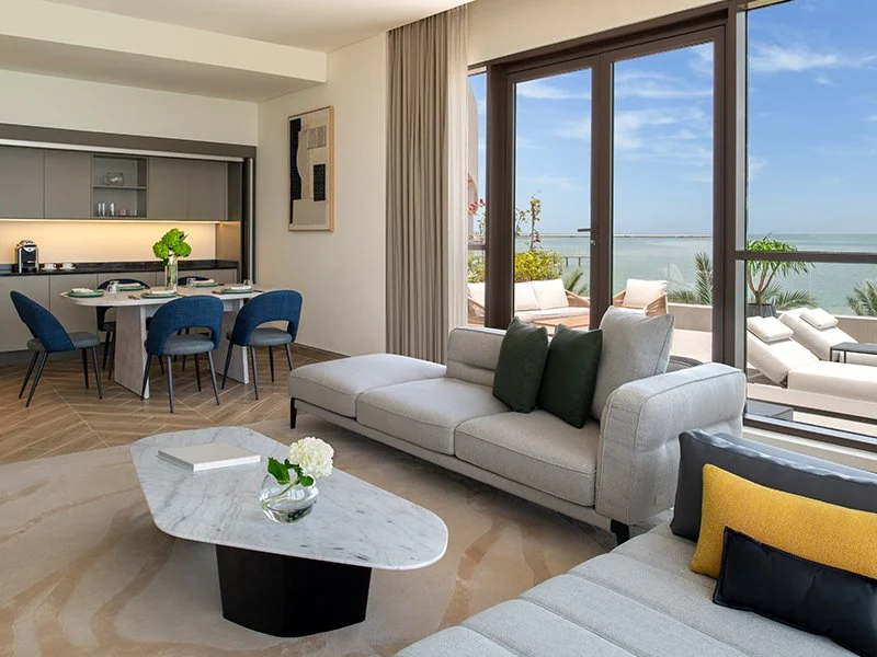 Living room with large sliding glass doors opening to balcony overlooking the ocean, modern furniture, and a dining table in the background.