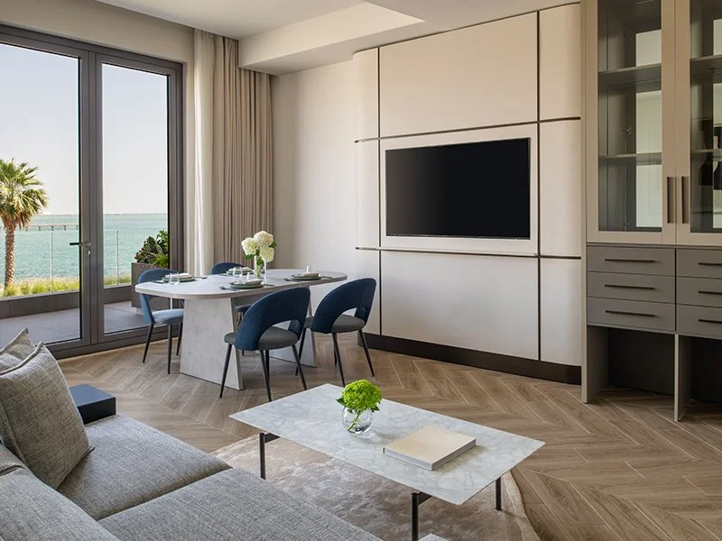 Modern living room with dining area, large sliding glass door opening to a balcony with ocean view, and contemporary decor including a gray sofa, a white coffee table with a small plant and book, and a wall-mounted TV.