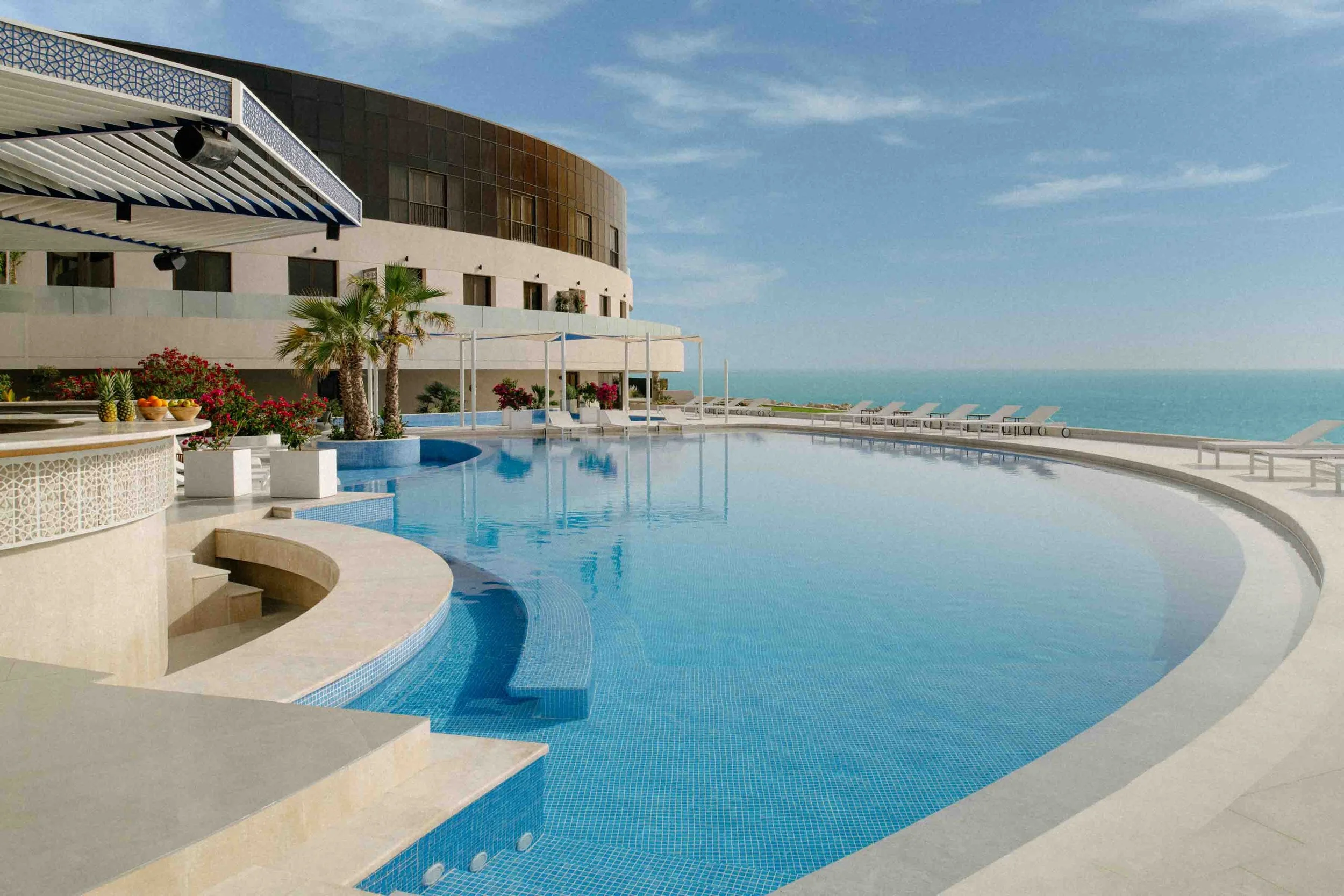 Luxury hotel rooftop pool area overlooking the ocean with lounge chairs, umbrellas, and tropical plants.