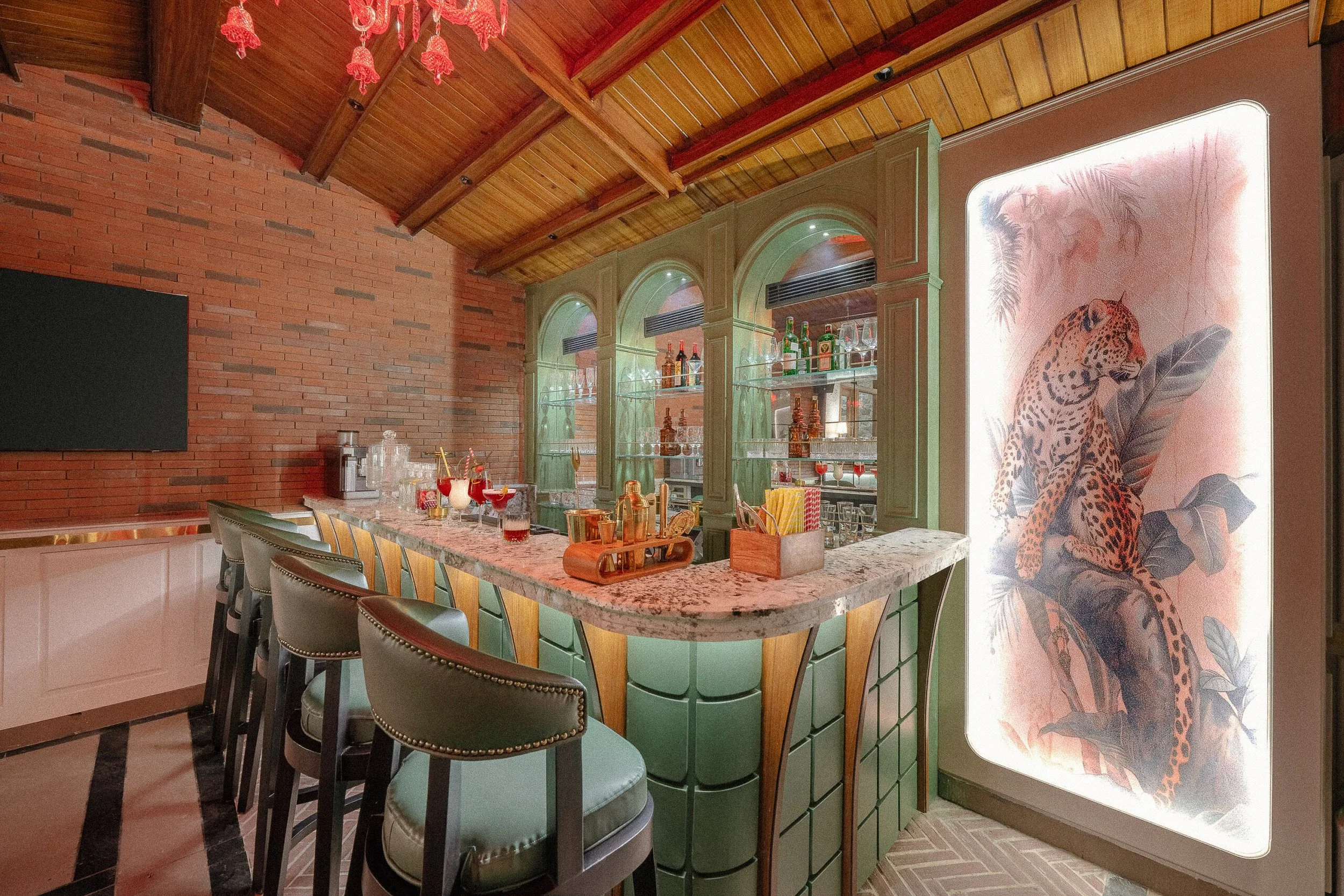 A bar area with a marble countertop, bar stools, green cabinets, and a wooden ceiling. There is a large illuminated painting of a cheetah on the right side and a brick wall on the left.
