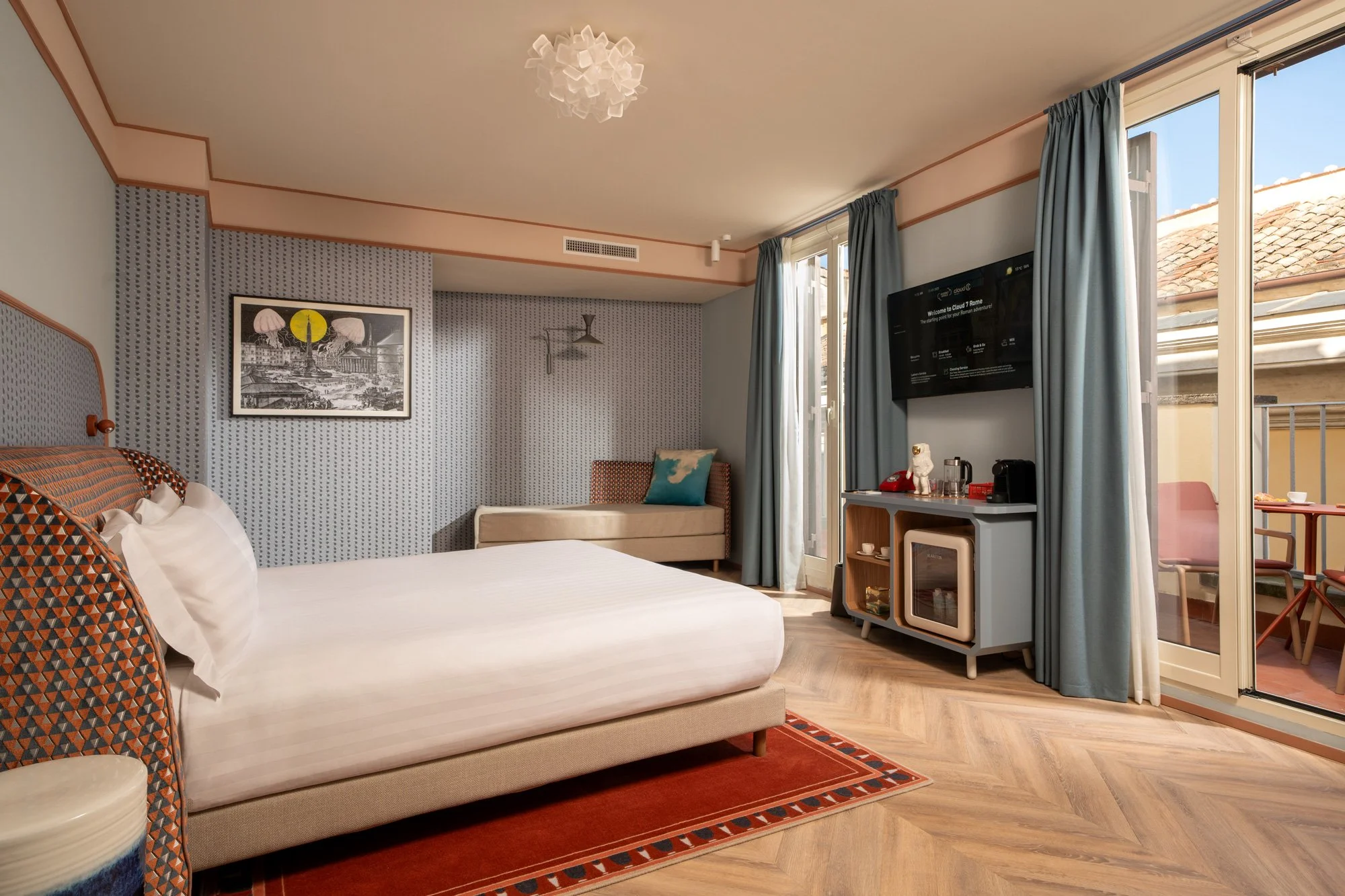A hotel room with a white bed, patterned pillows, a framed artwork on the wall, a beige sofa with a blue pillow, a flat-screen TV mounted on the wall, and balcony doors with blue curtains opening to a view of rooftops and a clear blue sky.