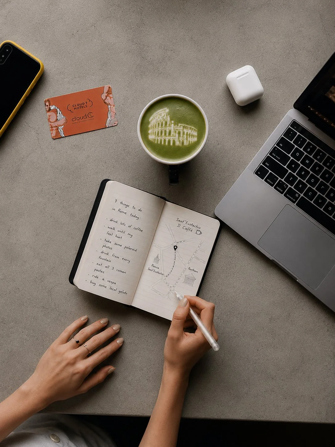 A workspace with a laptop, a cup of green matcha with a white illustration of the Colosseum, a closed white AirPods case, a yellow smartphone, a red hotel card, and an open planner with handwritten to-do list and a map of Rome. A person is writing or drawing in the planner.