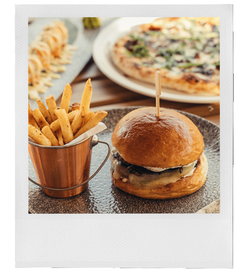 A cheeseburger with a wooden skewer, French fries in a metal cup, and a pizza in the background on a wooden table.
