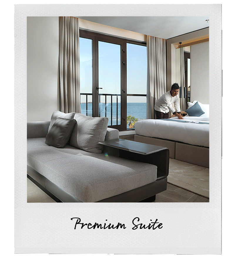 A hotel room with a seating area and a bedroom, featuring large windows with a view of the ocean, a person making the bed, and modern decor.