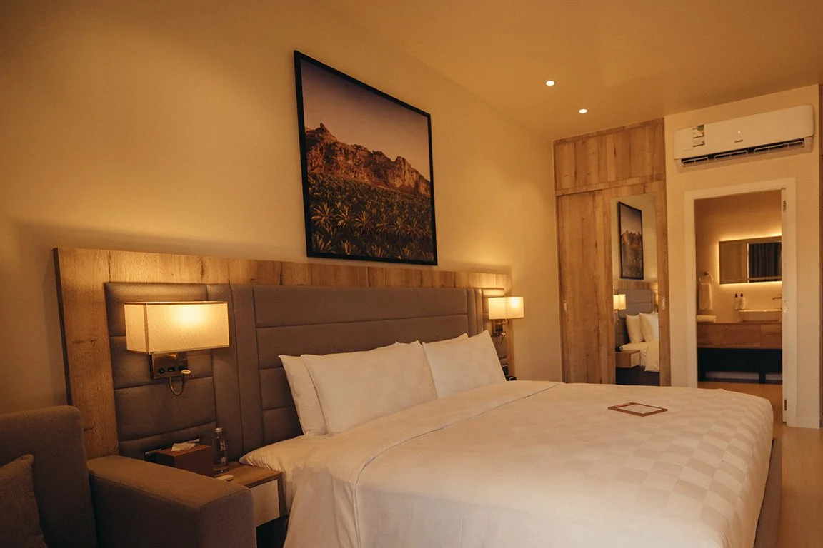 Hotel room featuring a large bed with white bedding, nightstands with lamps, artwork of a mountain landscape above the bed, a wooden closet, and a bathroom visible through an open door.