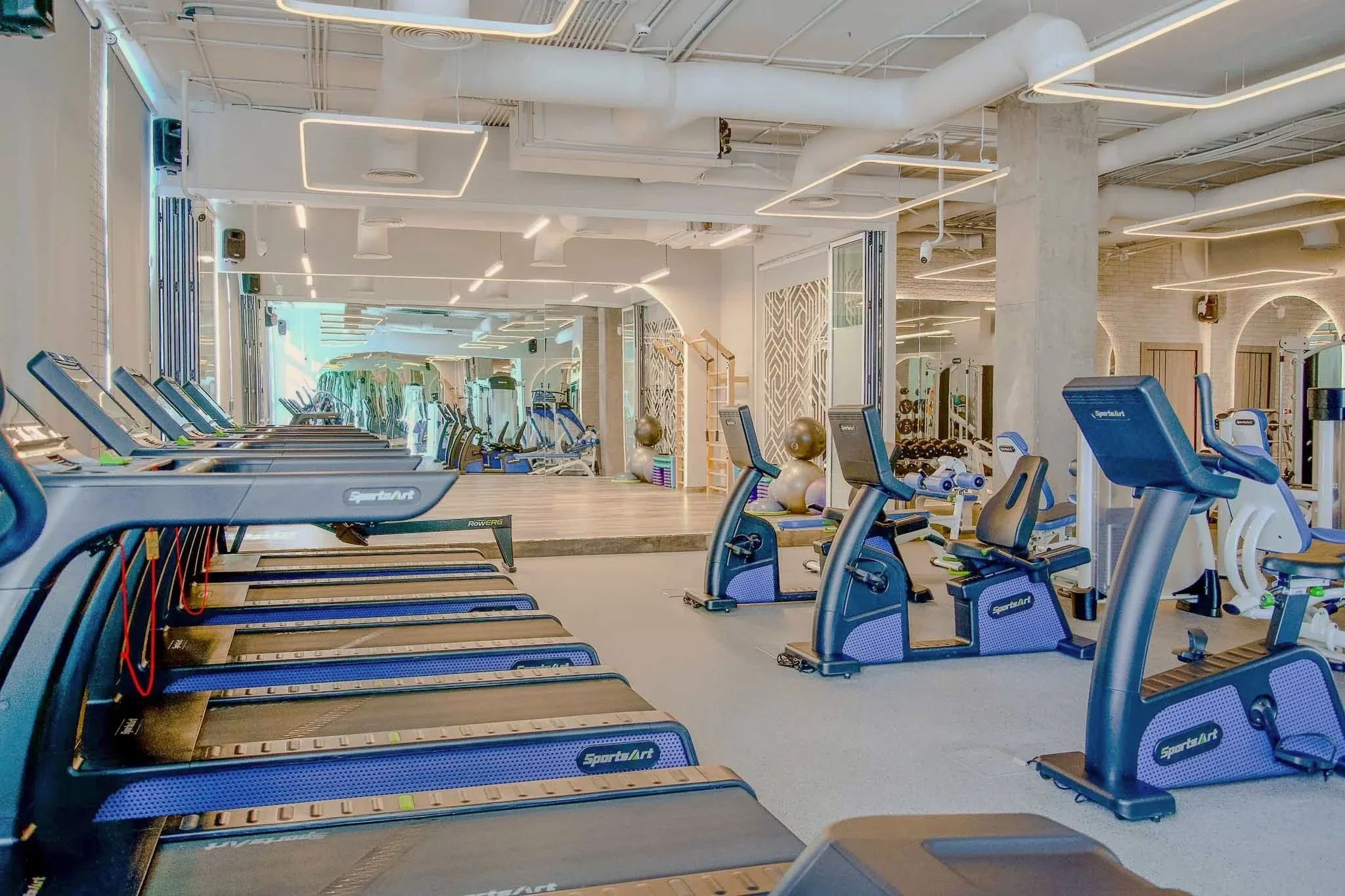 Empty gym with treadmills, exercise balls, weight racks, and mirrors, featuring modern lighting and industrial ceiling design.