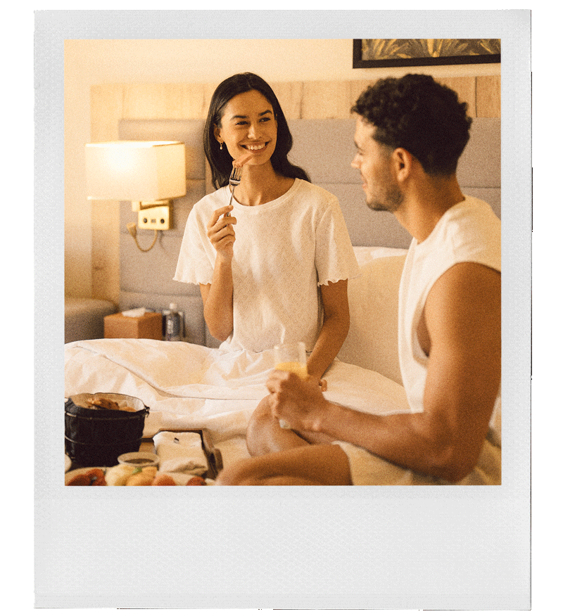 امرأة ورجل جالسان على السرير، يبتسمان، حيث تتناول المرأة طعامها بينما يحمل الرجل كوبًا من عصير البرتقال، وذلك في غرفة نوم مريحة تحتوي على طاولة بجانب السرير ومصباح.