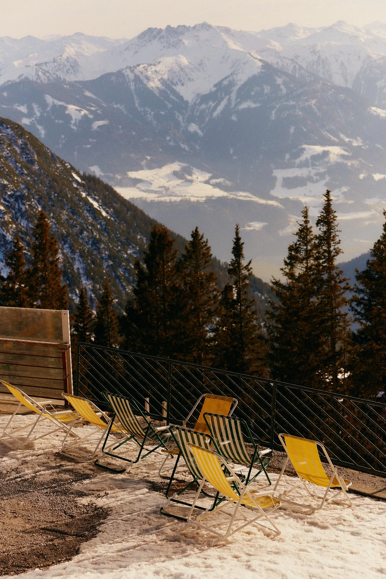 Multiple yellow, green, and striped lounge chairs on a snow-covered outdoor terrace with a mountain range of snow-capped peaks and evergreen trees in the background.