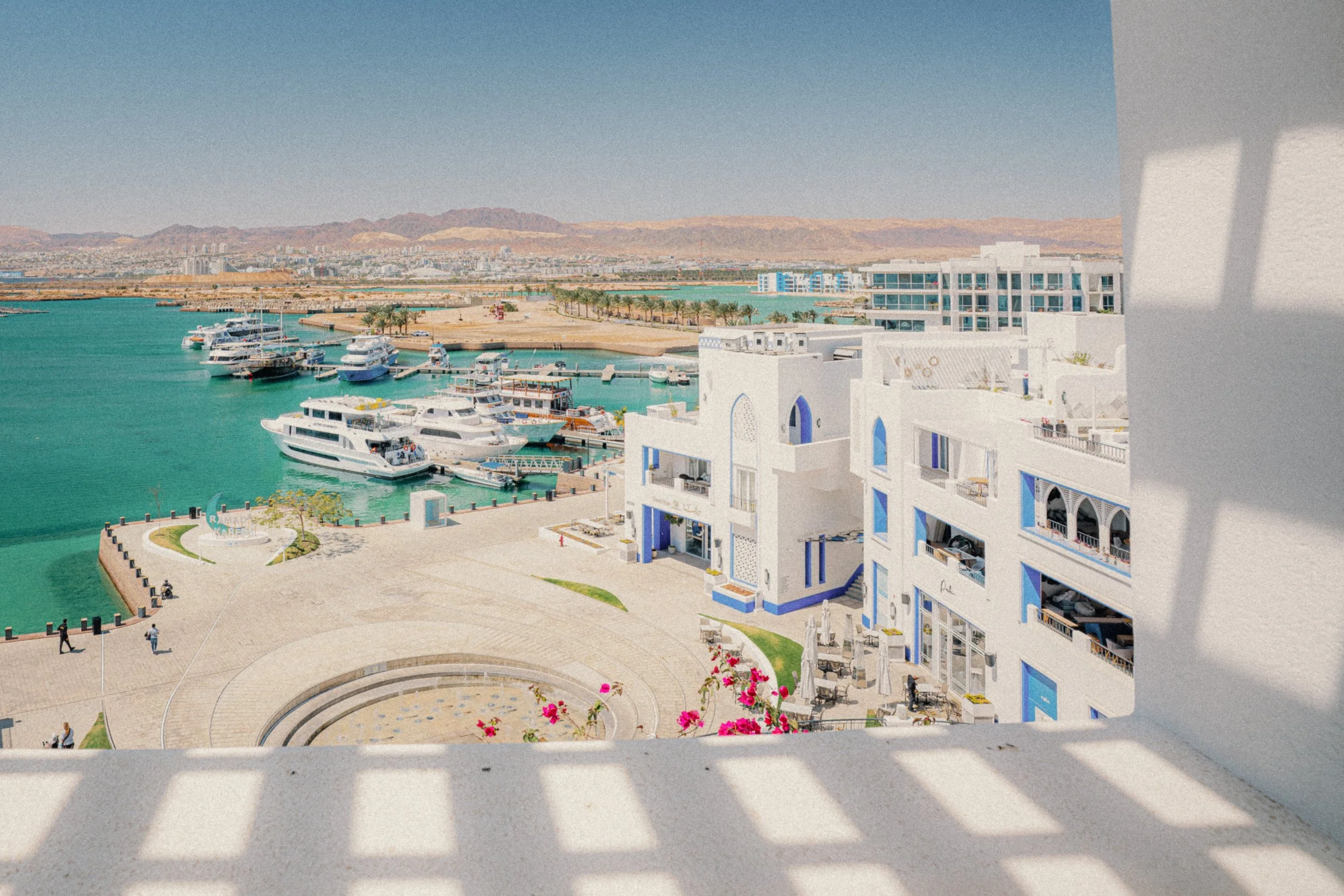 View of a marina with boats, a promenade, white buildings, and distant mountains, from a balcony with shadows on the railing.