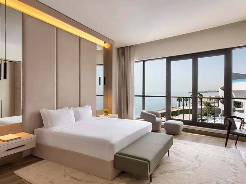 Modern hotel room with large window view of the ocean, a bed with white bedding, a beige upholstered headboard, a bench at the foot of the bed, side tables with lamps, and seating area with chairs and a small round table.