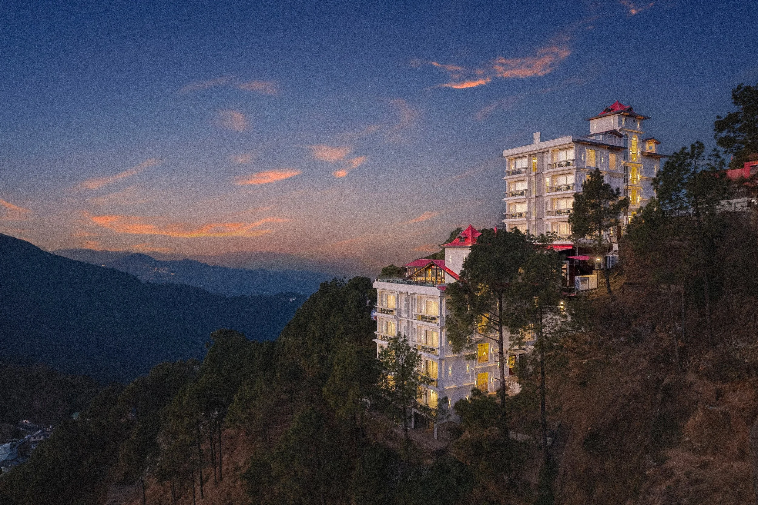 A multi-story white building with red roofs on a hillside, with trees and mountains in the distance during sunset or dusk.