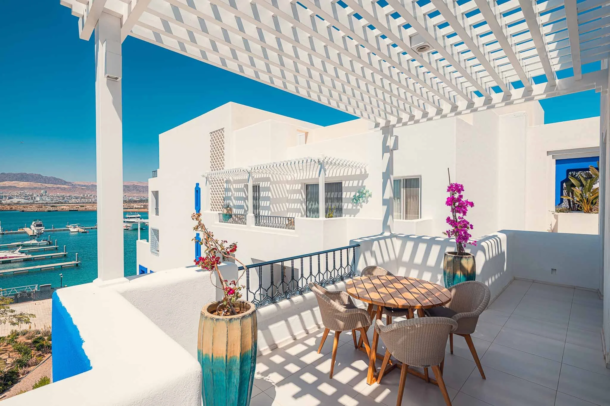 A balcony with a round wooden table and four wicker chairs, overlooking a marina with boats and a cityscape in the distance, under a white pergola, with white walls and colorful plants.