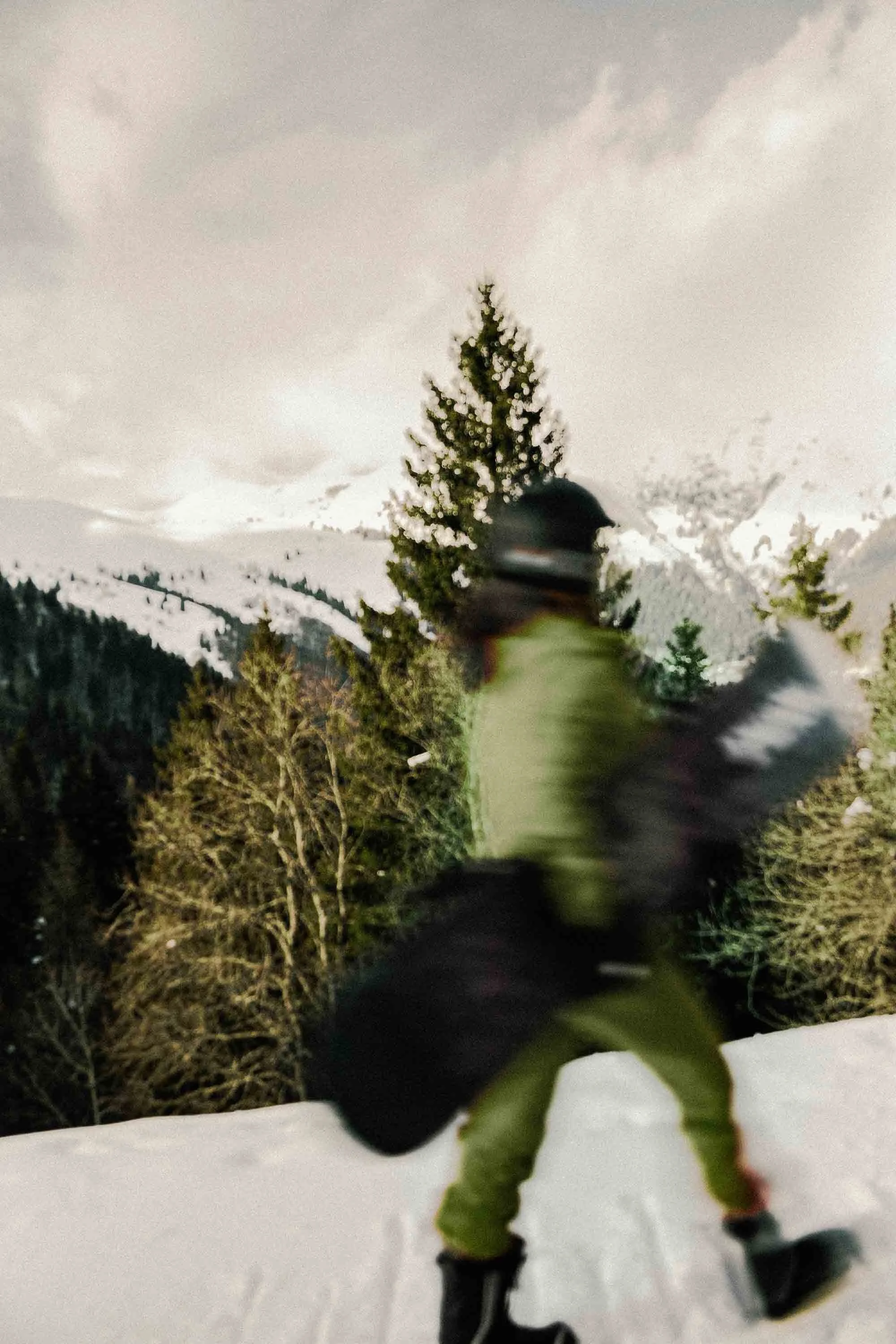 A person wearing a helmet and winter gear walking through a snowy landscape with pine trees and mountain in the background.