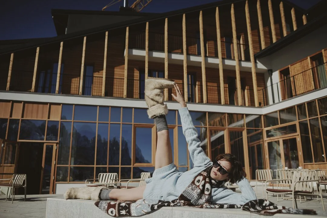 A woman in sunglasses lounging on a hotel balcony, stretching her leg upward, with a modern wooden building in the background.