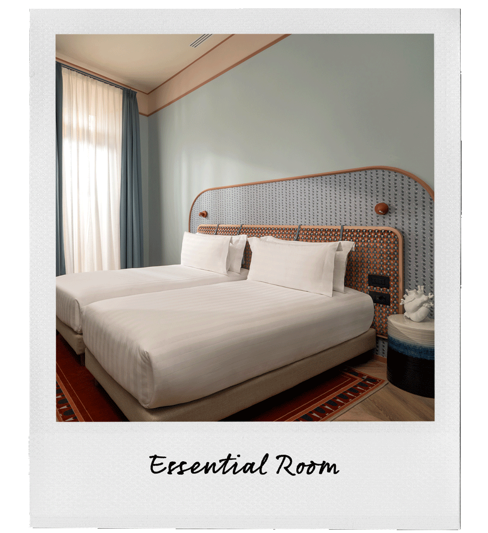 A hotel room with two single beds, white bedding, and pillows, next to a nightstand with a decorative sculpture, a patterned headboard, and a window with light-colored curtains.