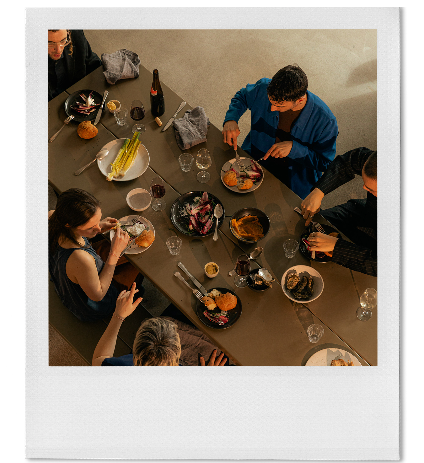 A top-down view of people dining at a long table with various plates of food, wine glasses, and utensils.