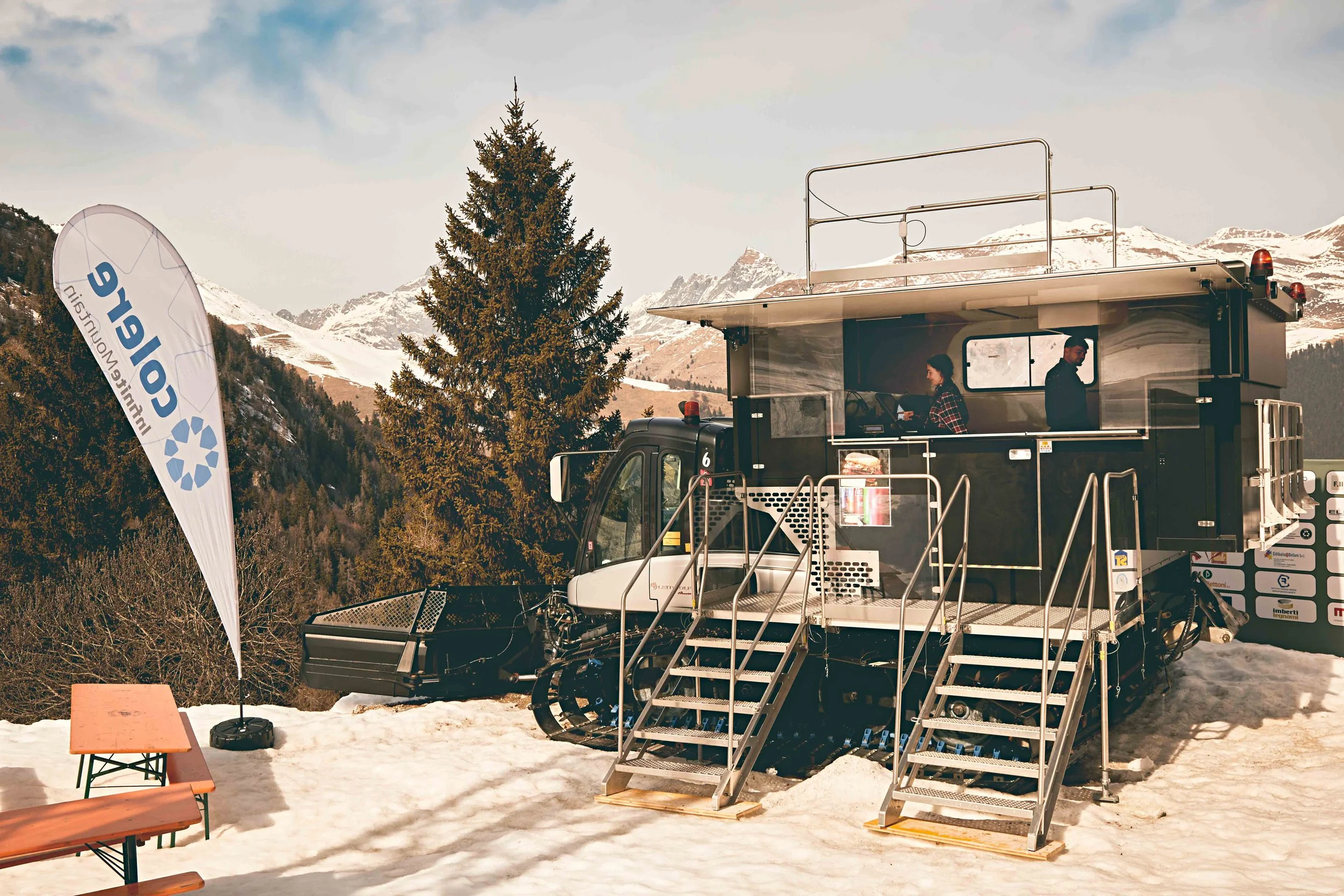 A snow-covered outdoor scene with a large ski lift tram, two people inside, a 'CEOLI' branded flag, benches, and mountains in the background.